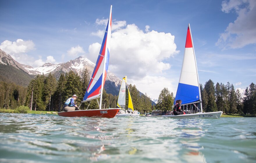 Segelboote auf dem Heidsee mit schneebedeckten Bergen im Hintergrund und zwei Personen beim Segeln auf dem klaren Wasser. | © Sundroina Pictures